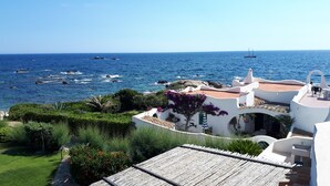 A coastal property with a view of the sea, featuring a white building with a terracotta roof, a garden with red flowers, and a wooden deck.
