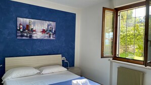 A bedroom with a bed, a ceiling fan, a window with a view of greenery, and a painting on the wall.