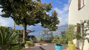 A courtyard with tiled flooring, potted plants, a tree, and a view of a lake and mountains.