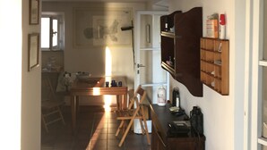 Un corridoio stretto con pavimento piastrellato, un tavolo di legno con sedie, una scrivania con un telefono e una mensola a muro con diversi oggetti.
