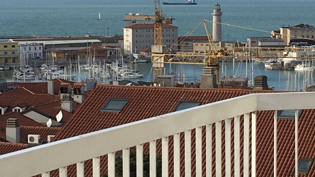 Un paysage urbain côtier avec un port de plaisance, des bâtiments aux toits recouverts de tuiles rouges et une grue.