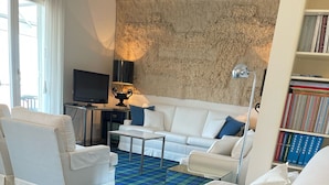 Une chambre d’hôtel avec un canapé blanc, un fauteuil blanc, une petite table, un téléviseur, une bibliothèque et un tapis à motifs écossais.