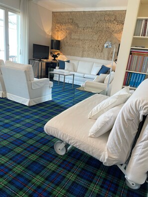 Habitación de hotel con un sofá blanco, un sillón blanco, una mesita, un televisor, una estantería y una alfombra estampada de cuadros.