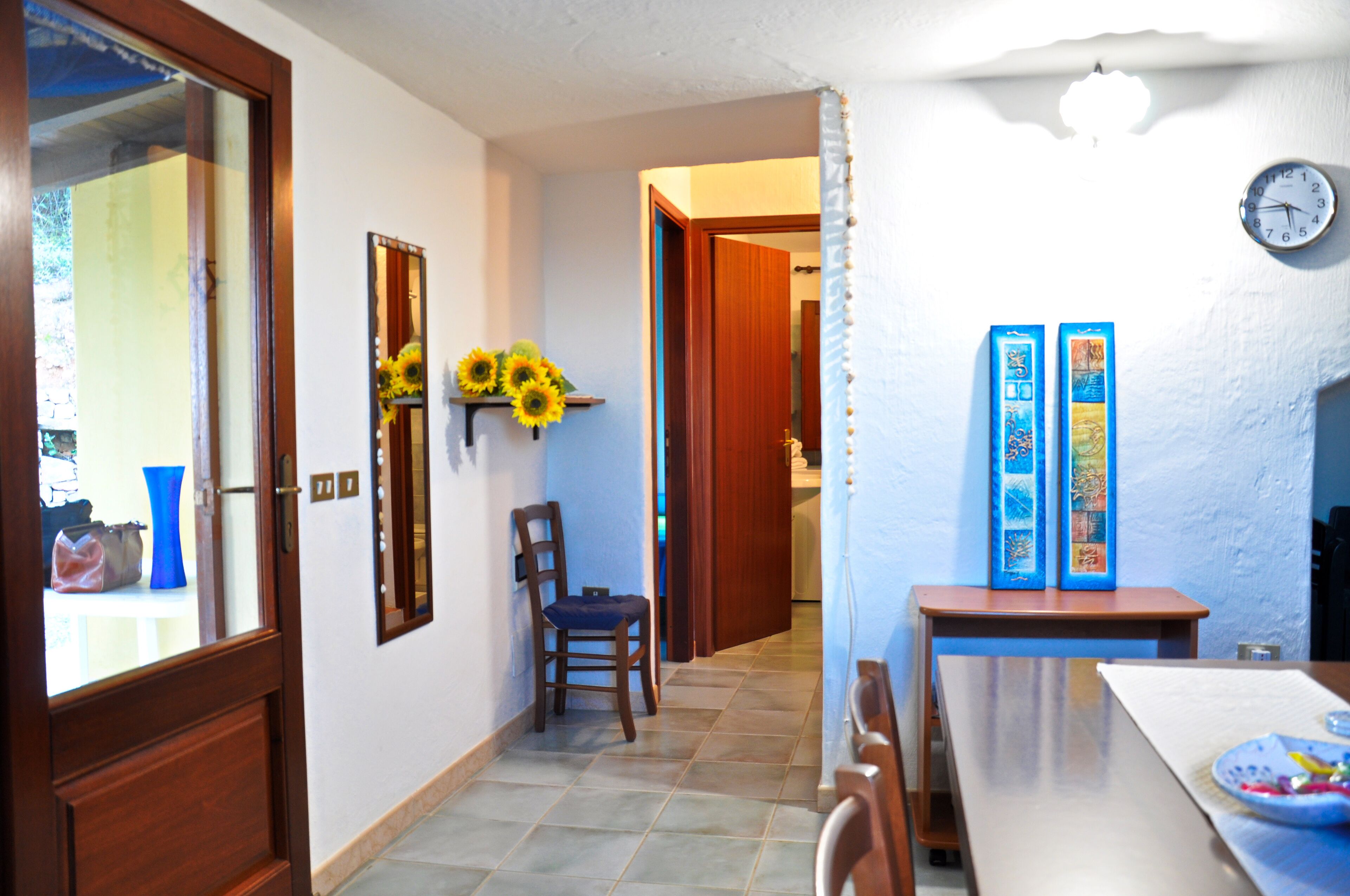 A hallway with a wooden door, a chair, a table with a plate, and a wall clock.