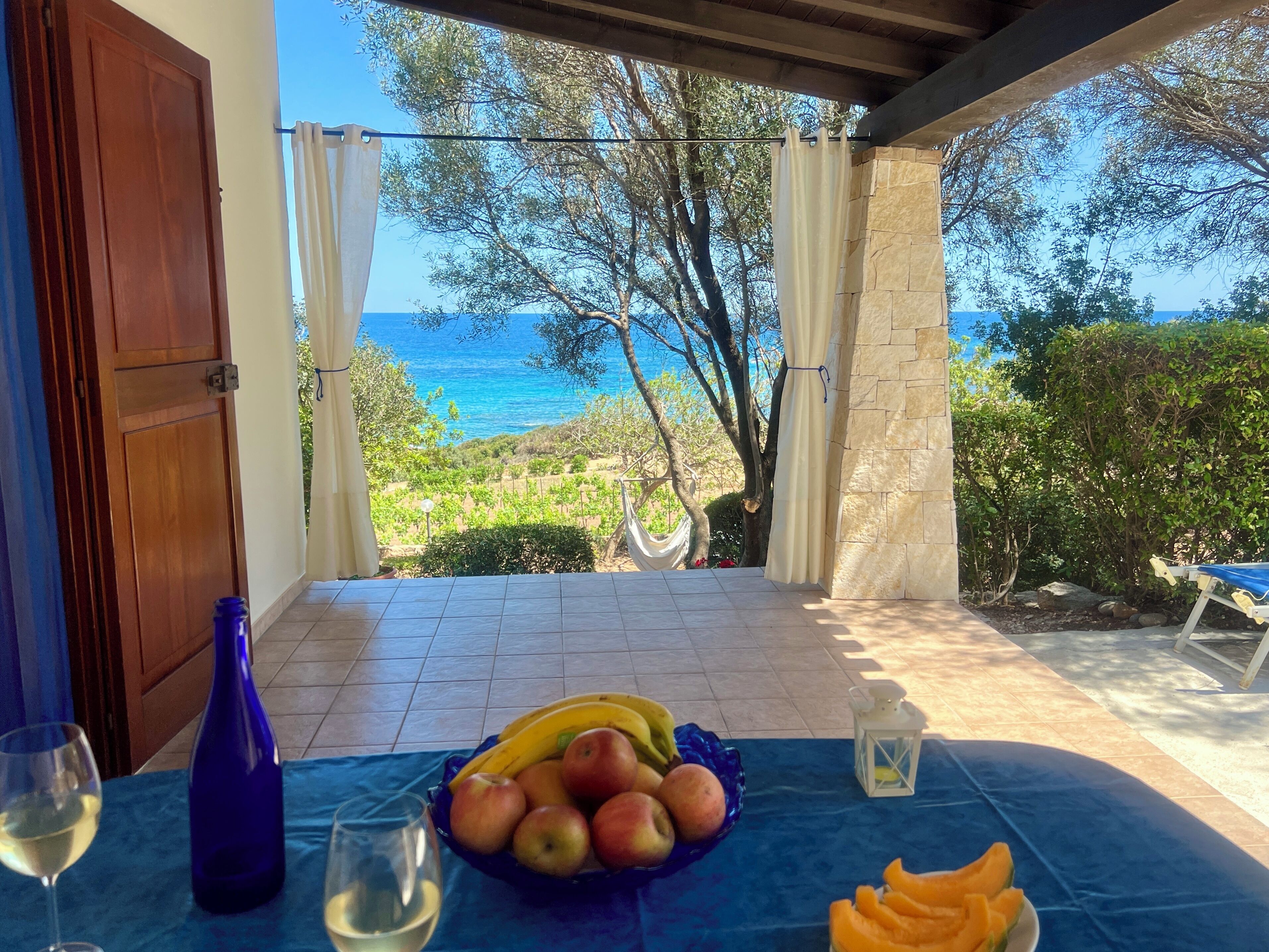 A terrace with a table set for a meal, a bottle, and a glass of wine, overlooking a scenic view of the sea and greenery.