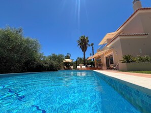 A swimming pool with a clear blue water surface, surrounded by lush greenery and a building with a tiled roof. - Large detached three bedroom villa with heated infinity pool and BBQ area. (Carvoeiro)