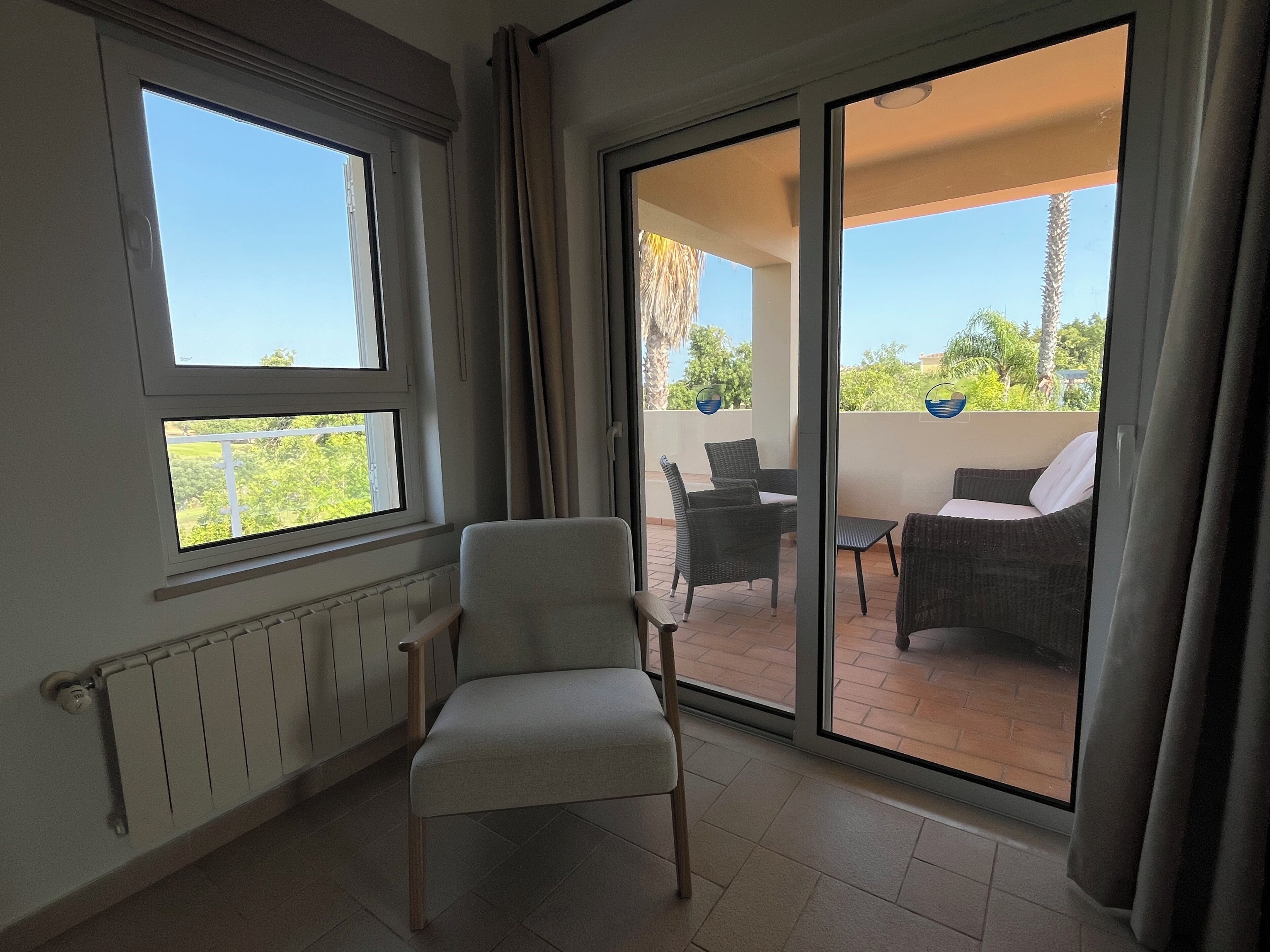 Een kamer met een stoel, een tafel en uitzicht op een balkon met rieten meubels en planten.