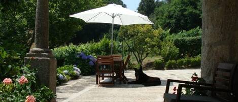 A covered patio area with a stone floor, a wooden bench, a table, and an umbrella providing shade.