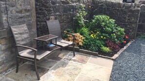 A patio area with a stone wall, a metal chair, a small table, and a garden bed with various plants and flowers. - Fern House, Grassington; central yet parking & quiet, light & period elegance (Grassington North Yorkshire)
