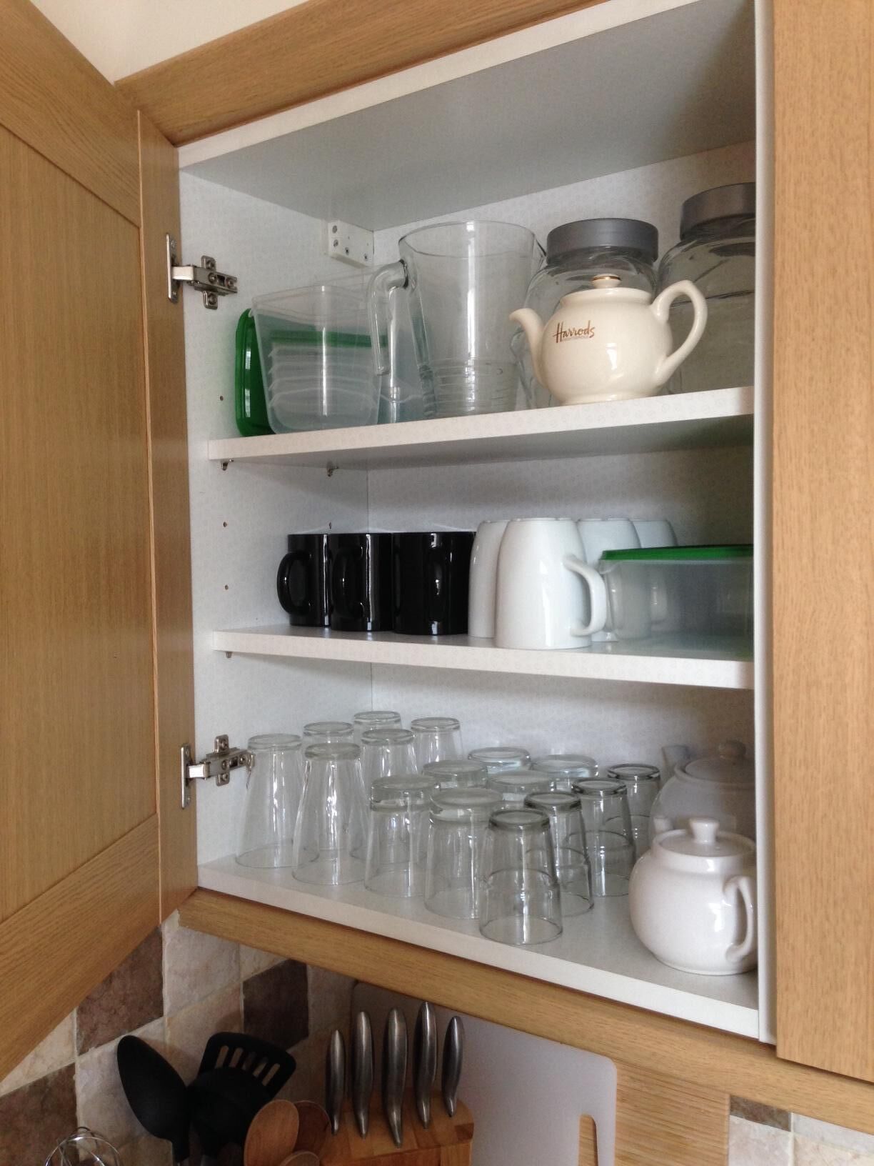 A kitchen cabinet with shelves holding various glassware, including tumblers, mugs, and a teapot.