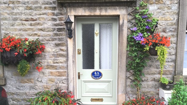 A stone building with a green door, a blue sign with '4A', and various potted plants around the entrance. - Fern House, Grassington; central yet parking & quiet, light & period elegance (Grassington North Yorkshire)
