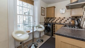 A compact kitchen with a washing machine, a gas stove, and a small dining area with a glass table and white chairs. - Fern House, Grassington; central yet parking & quiet, light & period elegance (Grassington North Yorkshire)