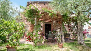 A rustic stone building with a wooden pergola covered in flowering vines, outdoor seating, and potted plants.