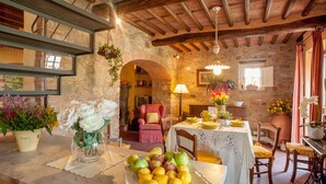 A rustic dining room with a stone wall, wooden beams, a table set with fruits, and a chair.