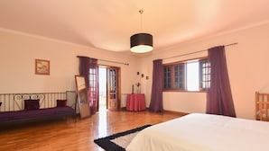 A bedroom with a bed, a sofa, a window with curtains, a door, and a hanging light fixture.