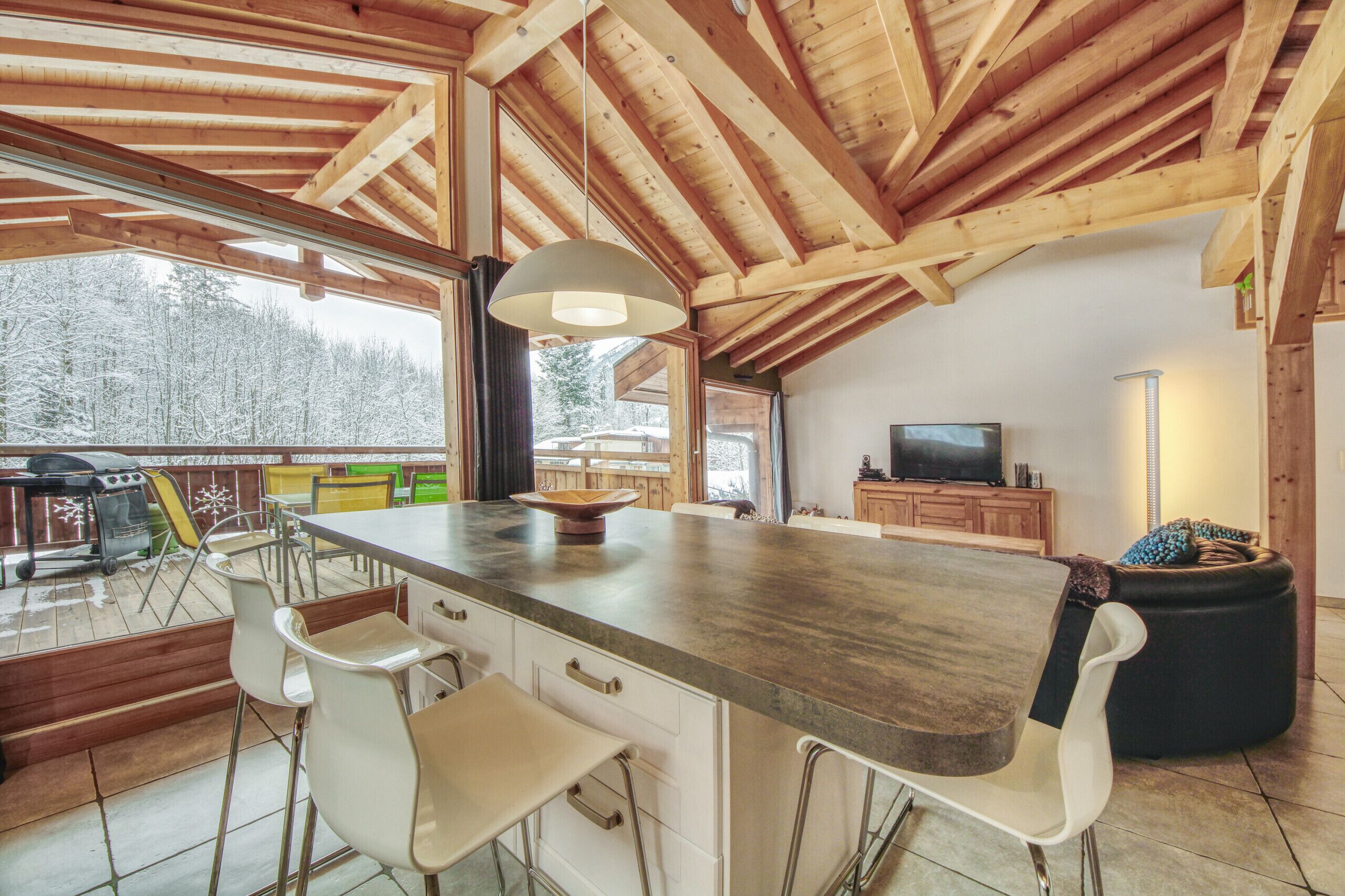 A kitchen with a wooden ceiling, a large island with bar stools, a television, and a view of a snowy landscape.