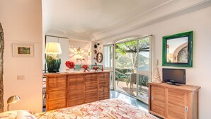 A bedroom with a bed, wooden dresser, TV, and a balcony with a view.