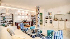 A modern living room with a sofa, coffee table, and patterned chairs. A kitchen area with white cabinets and a tiled backsplash is visible in the background.