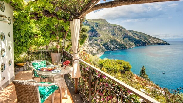 A balcony with wicker furniture, a table with a plate of food, and a view of a coastal landscape with cliffs and the sea.