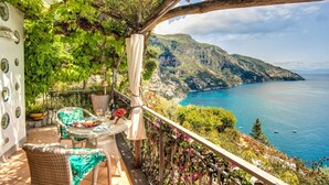 A balcony with wicker furniture, a table with a plate of food, and a view of a coastal landscape with cliffs and the sea.