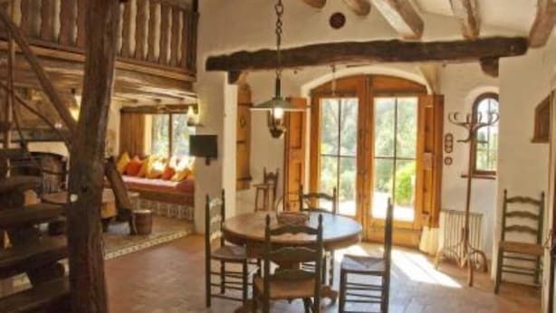 Un intérieur rustique avec des poutres apparentes, une table ronde en bois entourée de chaises et une grande fenêtre en arc qui laisse entrer la lumière naturelle.