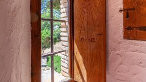 Une porte en bois ornée d’un motif solaire et sur laquelle est gravé le mot « BACH », à côté d’une fenêtre donnant sur un paysage verdoyant et un mur de pierres.