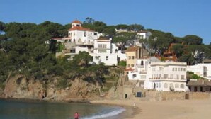 Une zone côtière avec des bâtiments construits sur une colline, une plage où se trouvent des gens, et un ciel dégagé.