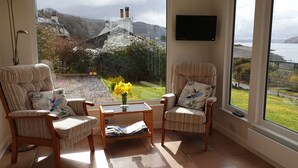 A cozy room with two striped armchairs, a small table with a vase of yellow flowers, and a view of a lake and mountains through large windows.