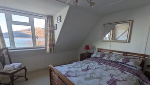 A bedroom with a bed, a chair, a nightstand, a lamp, a mirror, and a window with a view of the sea.