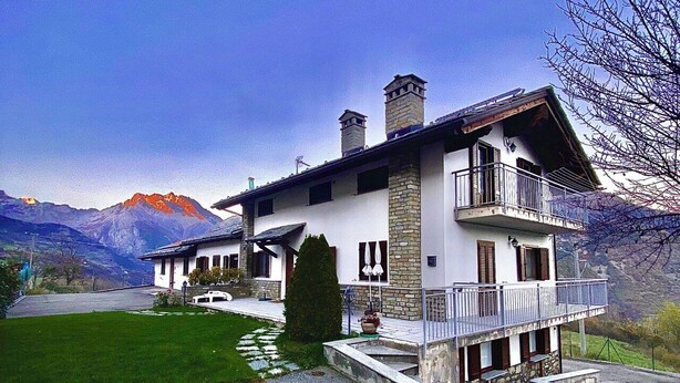 Una casa de dos pisos con balcón, chimenea de piedra y montañas al fondo.