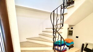 Una escalera con peldaños color beige y barandilla de metal, que lleva a un rellano con una pequeña mesa y ropa doblada.