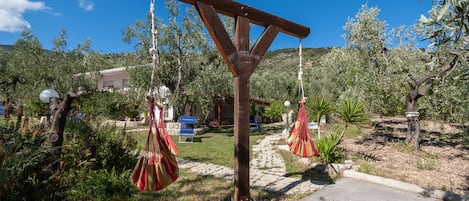 Une pergola en bois, ornée de hamacs rayés de rouge et de blanc, est installée dans un jardin où l’on trouve un sentier en pierre et des oliviers.