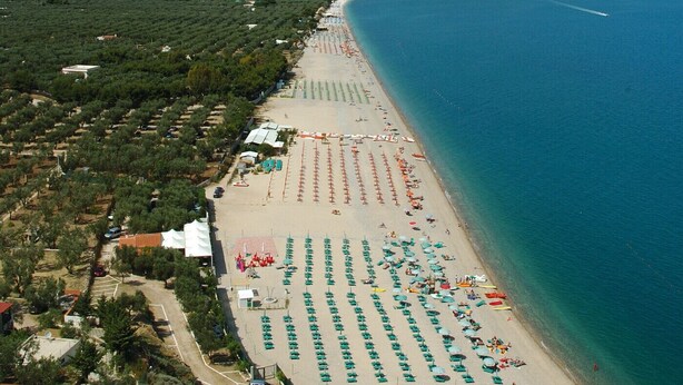 Une plage côtière bordée de rangées de parasols et de chaises longues, une mer bleue et limpide, et un paysage vallonné en arrière-plan.