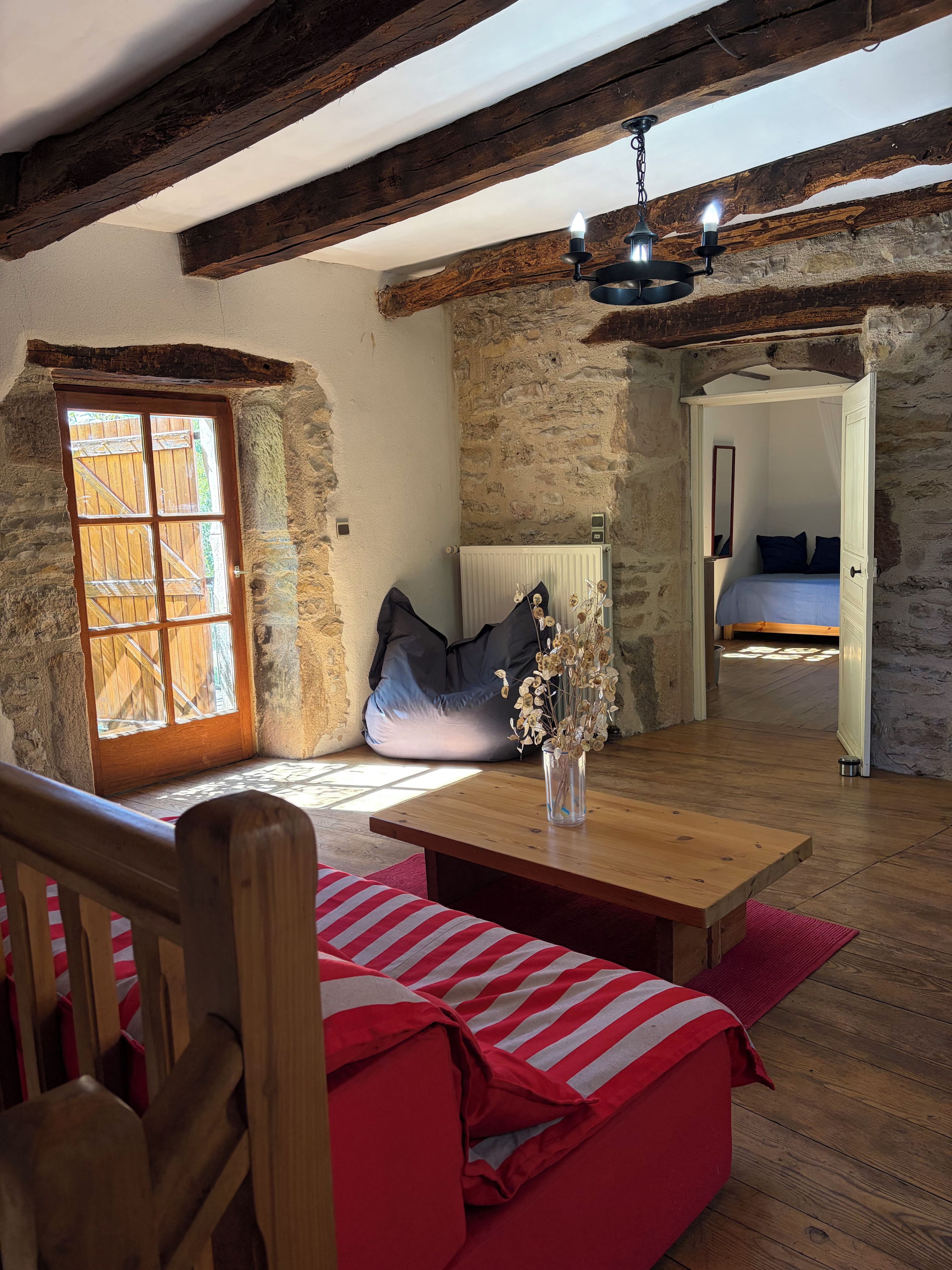 Una habitación rústica con un banco de madera, una mesa con un florero, una pared de piedra y una cama con colcha azul.