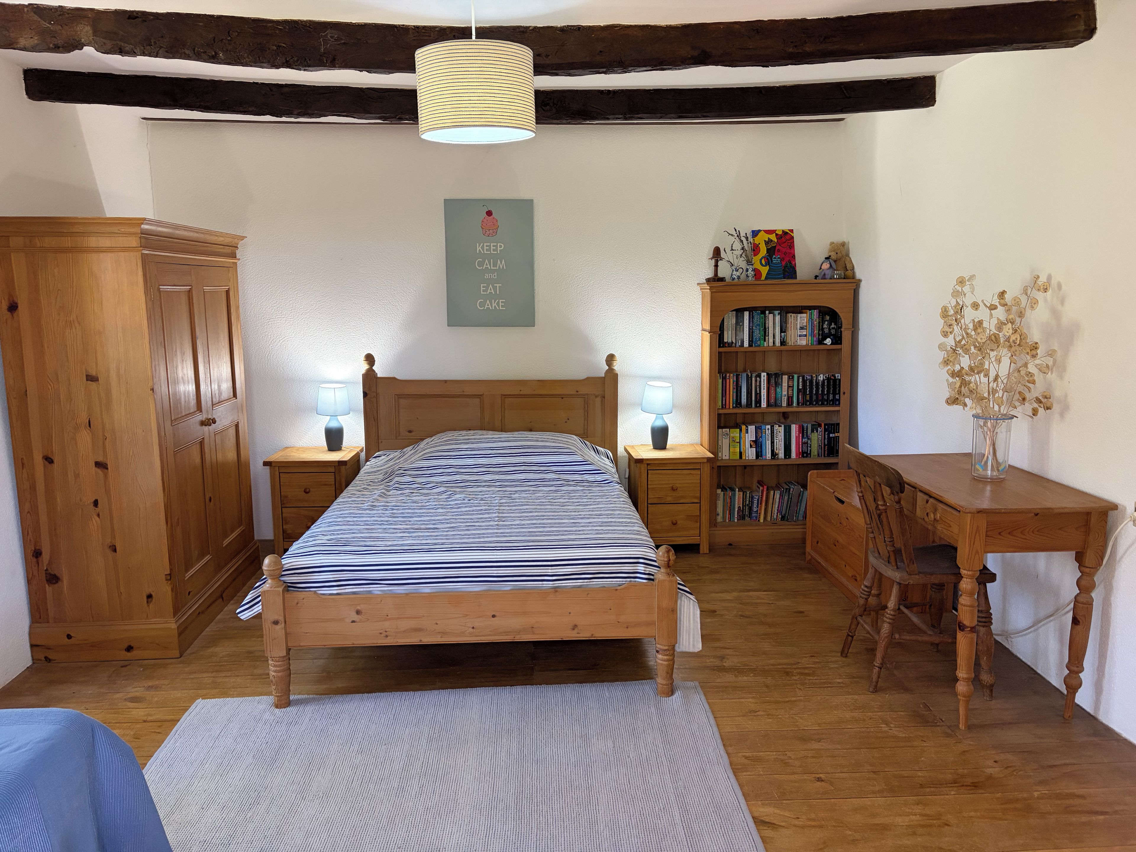 Une chambre à coucher avec un lit en bois, des tables de chevet avec des lampes, une bibliothèque et un bureau agrémenté d’un vase de fleurs séchées.