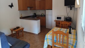 Een kleine keuken met houten kastjes, een magnetron, een koelkast en een eethoek met een tafel en stoelen.
