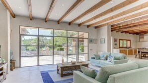 A spacious living area with a large sliding glass door leading to an outdoor patio, a kitchen area with wooden beams, and a seating area with light green sofas.