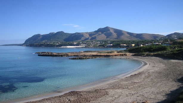 Una playa costera con aguas transparentes, afloramientos rocosos y una montaña al fondo.
