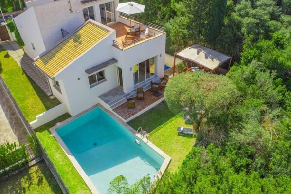 Una casa moderna de dos pisos con piscina, un patio cubierto y un exuberante jardín verde.