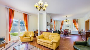 Un salon spacieux avec un canapé jaune, une table basse en verre et un fauteuil vert. On y trouve également deux autres fauteuils jaunes, un coin repas avec une table et des chaises, ainsi qu’une cuisine équipée d’une cuisinière et de placards.
