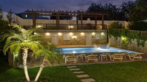 Une piscine dotée d’une terrasse en bois et de chaises longues, entourée de murs en pierre et de palmiers.
