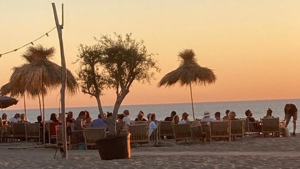 Un área de estar frente a la playa con sillas de madera y sombrillas de paja, con vista al océano.