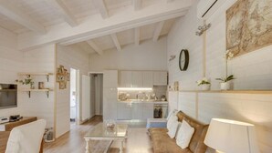 Un salon moderne avec un canapé, une table basse et une lampe. On aperçoit au fond une cuisine avec des meubles blancs et un coin repas comprenant une table et des chaises.