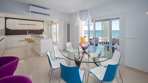 Une cuisine moderne dotée d’un îlot central, un coin repas avec une table en verre et des chaises bleues, et un balcon offrant une vue sur la mer.