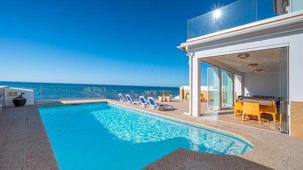 Une piscine à l’eau cristalline, entourée de chaises longues et dotée d’un patio vitré offrant une vue sur l’océan.