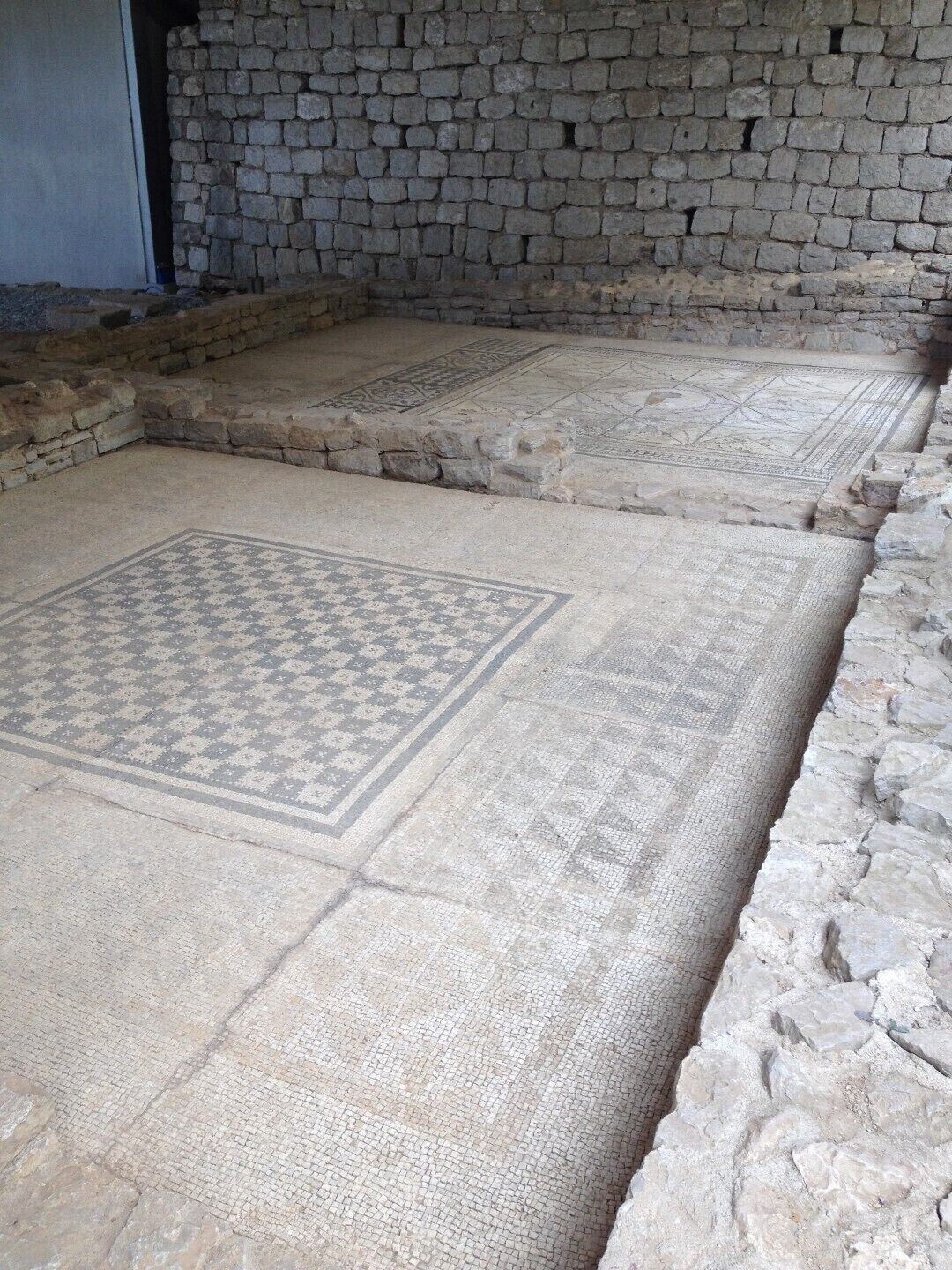 Un sol en pierre orné de motifs complexes en mosaïque, un mur en pierre et une porte partiellement visible.