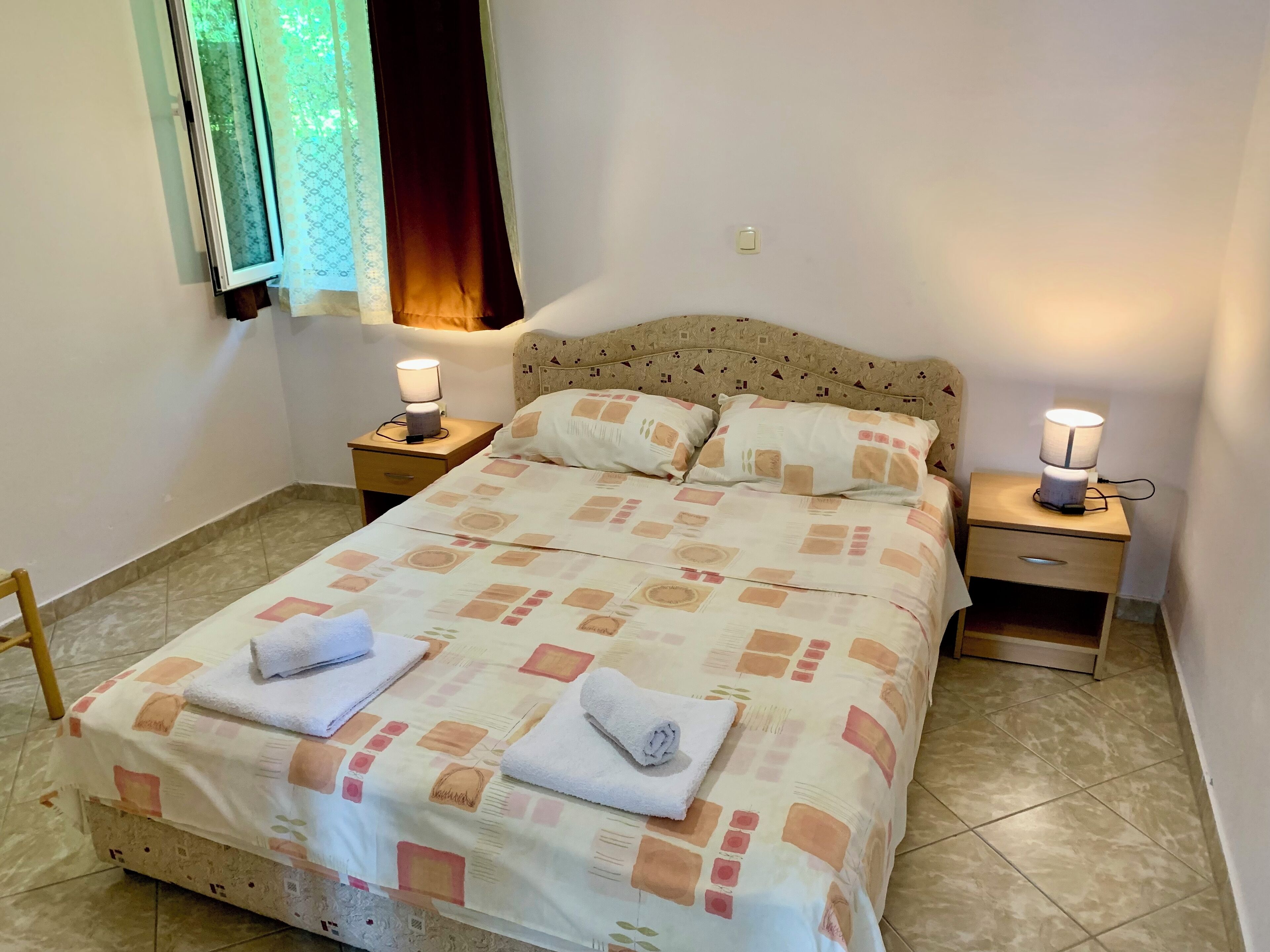 A neatly made bed with a patterned bedspread, two bedside lamps, and a window with curtains.