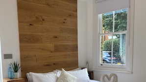 A bedroom with a wooden headboard, white bedding, a floral quilt, and a window with a view of the outside. - Beautiful One bedroom contemporary Interior Designed  Flat just off Kew Green (Kew Richmond)
