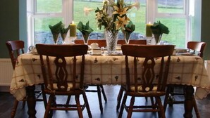 Une table à manger dressée pour quatre personnes, agrémentée d’un centre de table floral, de deux chaises et offrant une vue sur un champ verdoyant grâce à de grandes fenêtres.