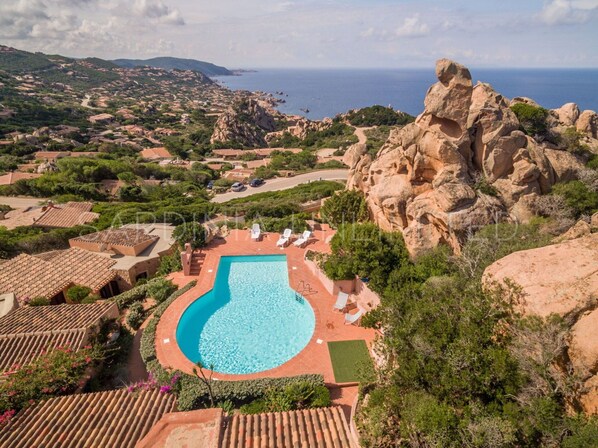 Uma grande piscina rodeada por um terraço com espreguiçadeiras, com vista para terreno rochoso e o litoral.
